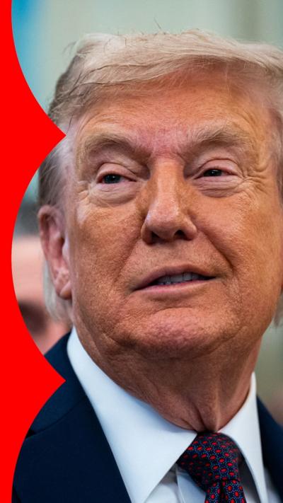Donald Trump with a slight smile, looking to the left. He wears a dark suit jacket, white shirt and blue tie with red dots