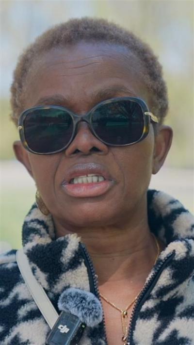 A woman with short hair and sunglasses speaks towards a camera off screen. She is wearing a printed sweater and a small microphone. The outdoor background is blurred behind her.