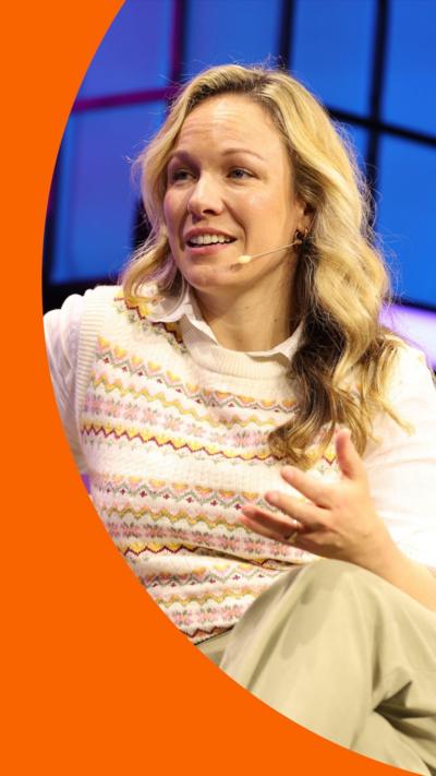 Eline Van Der Velden has long blonde hair. She is seated, and wearing a white shirt, sleeveless jumper and beige trousers.