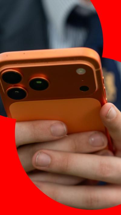 A close-up of a pupil's hands holding a metallic orange mobile phone. They are wearing a school blazer, shirt a