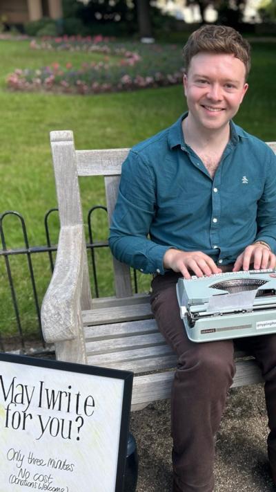 Man looking in to camera, sitting on a wooden bench. He's got brown hair, green shirt, with a typewriter on his lap. There's a green lawn behind him and bed of pink flowers. 