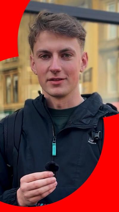 A man with short brown hair in a black jacket stands in the street 