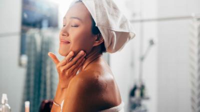 A woman stands in a bathroom with her eyes closed, touching her face. Her hair and body are wrapped in towels