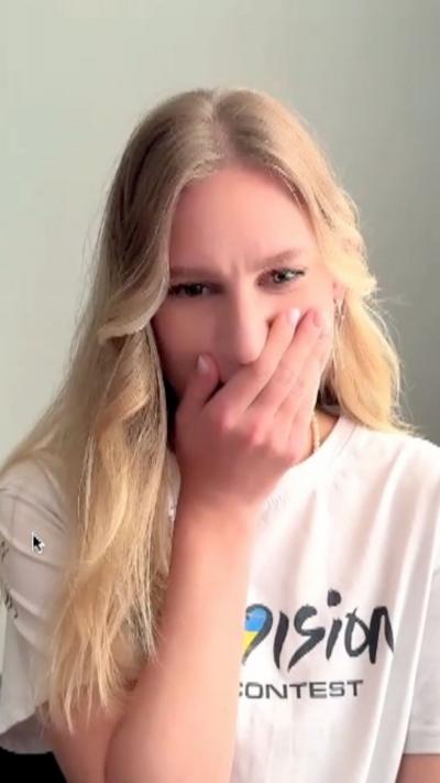 A woman, Tetiana, holds her hand over her mouth in shock. She has long blonde hair and wears a Eurovision T-shirt
