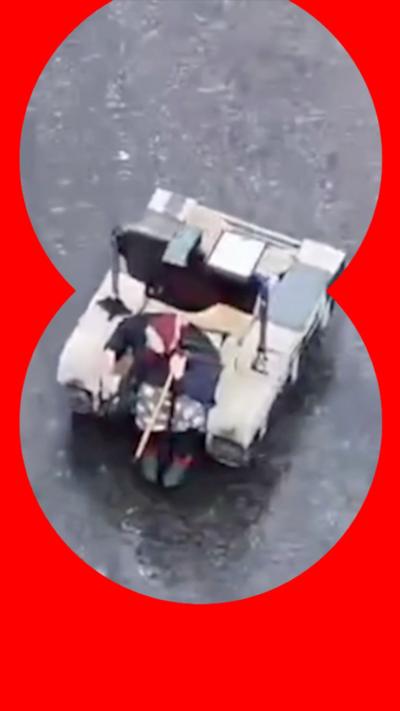 An older woman wearing a red scarf on her head sits on a square robot vehicle holding a walking stick.