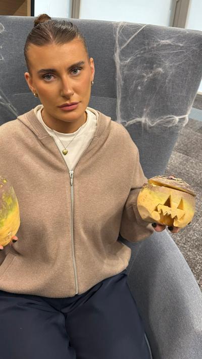 A woman sat on a grey chair in a beige cardigan. She is holding two turnips.
