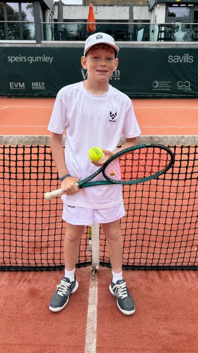 A young boy in tennis whites holding a racket and ball on a tennis court.