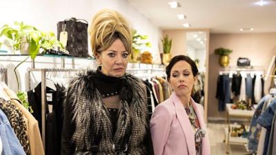Two women stand in a clothes shop - Mandy looks unsure, the salesperson looks in a judging manner