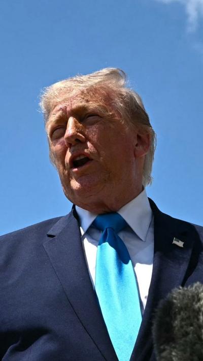 Donald Trump, wearing a blue suit and teal coloured tie, speaks to reporters (off camera) with a blue sky behind him.