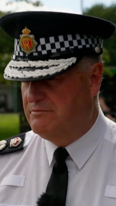 A white man with short grey hair in a police outfit including a shirt and tie and a police hat with the badge on the front