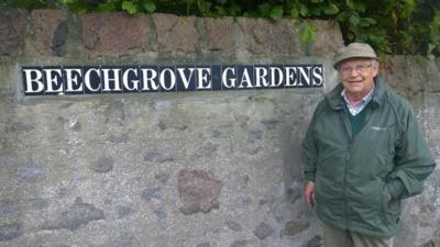 Jim McColl wears a tweed cap and green jacket and jumper with a checked shirt. He stands in front of a Beechgrove Gardens sign.