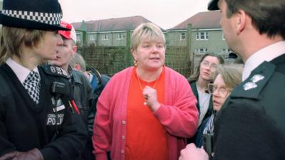 Margaret Haney photographed in 1997 outside a house on Stirling's infamous Raploch estate, with two police officers to either side during a 'paedophile-hunter' raid