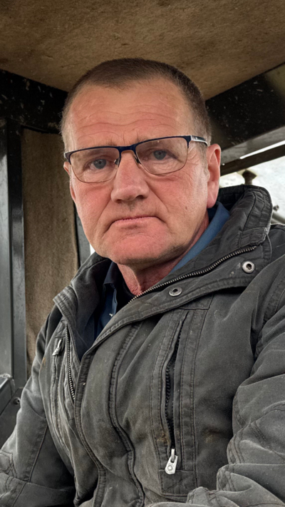 A farmer, a man with short black hair wearing glasses and a coat, sitting in the cab of a tractor on a rainy overcast day.