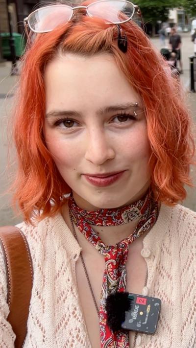 A young woman with shoulder length red hair looks at the camera. She is wearing a cream cardigan, and paisley scarf. She has a pair of glasses resting on top of her head.