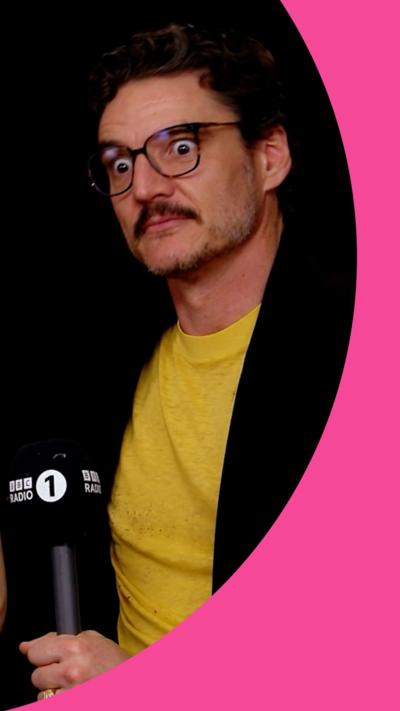 Pedro Pascal looks shocked while holding a Radio 1 mic. He wears a yellow T-shirt under a black jacket 