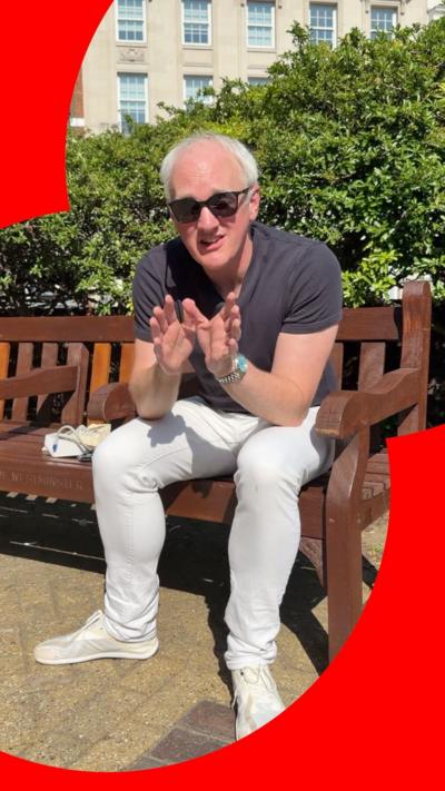 A man in white trousers and a grey tshirt and suglasses sits on a bench in the sun