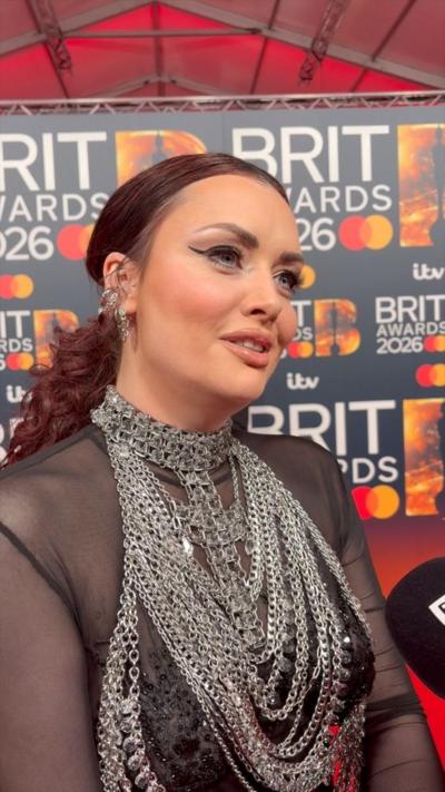 A woman on the red carpet, she has red hair tied back and a mesh top with huge chain necklaces.