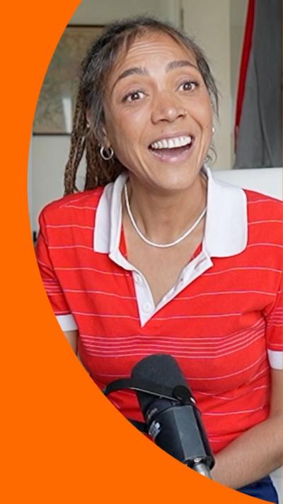 Miquita Oliver wears a red polo shirt and sits in a white office chair with a mic in front of her. She is smiling.