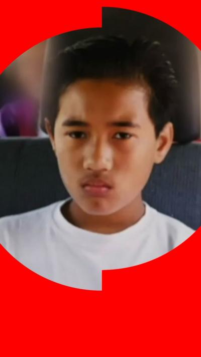 A young boy with short black hair sits in a motor vehicle. He is wearing a white T-shirt and looks upset.