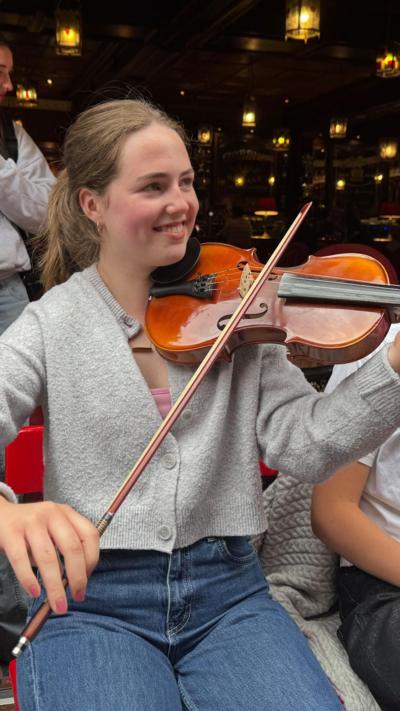 A woman smiling with a grey cardigan and blue jeans - playing a fiddle. 