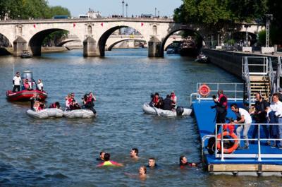 People seen swimming in the river Seine. There are also support boats in the river, and people gathered nearby on a floating pier.