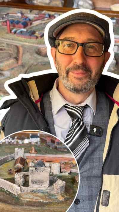 Steve stands inside a wooden shed wearing a winter jacket over formal clothing, beside a detailed model town set on top of cabinets. The shed’s interior is lined with wooden panels and shelves holding tools, boxes, and assorted materials.