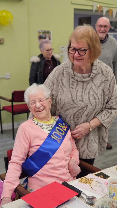 Smiling woman sat down at a table with a 100th birthday sash across her chest while another woman is standing beside her smiling too.