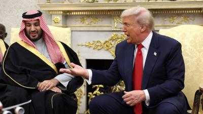 Two men sitting on gold chairs talking, one in a suit and one in Saudi attire