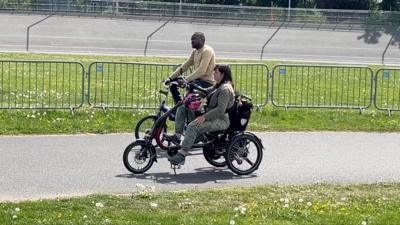 Two individuals ride specially adapted bicycles along a path lined with green grass and safety barriers.