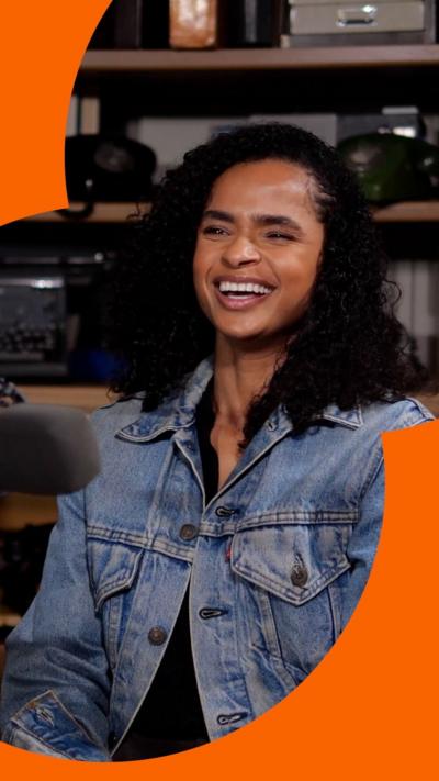 A woman with shoulder-length black curly hair laughing wearing a denim jacket.