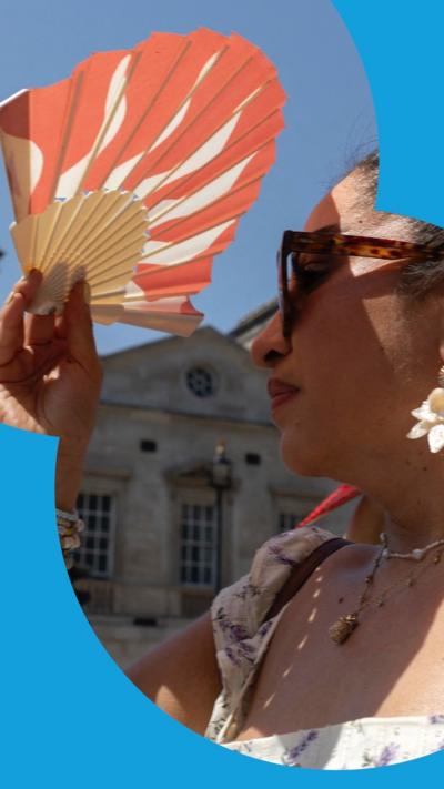 A woman with dark hair wears sunglasses and shades herself with an orange and white fan She is wearing a summery top, two necklaces and large flower earrings.