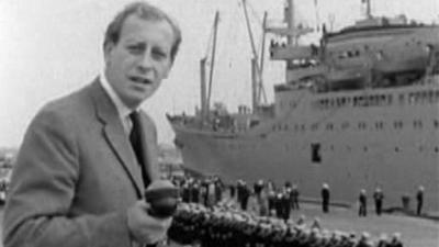 Black and white image of Peter Woods, standing on the left of the frame in a suit and tie, holding a microphone, looking down the lens. In the background, the Upshur ship is docked and the vessel's crew are lined up in uniform on the dockside.