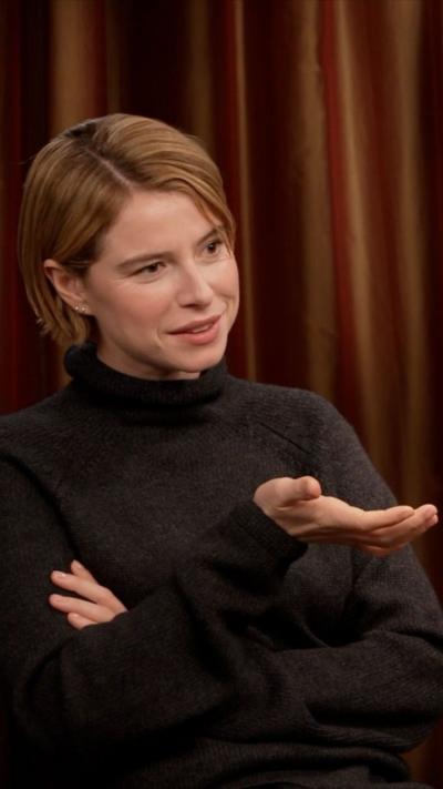 Jessie Buckley wears a dark grey jumper and sits with her arms crossed