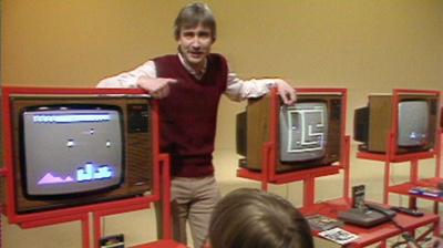 The presenter stands between two televisions showing video games