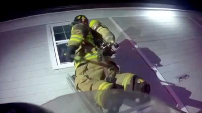 A firefighter kneels on ladder holding a hammer to a second-story window