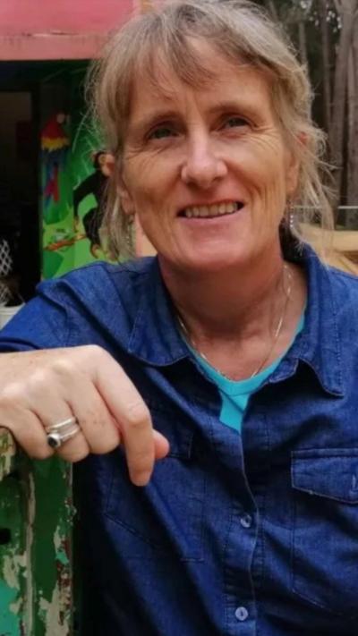 Gena Heraty, a woman with longish fair hair, smiling at the camera.  She has a fringe and the rest of her hair is loosely tied back. She is wearing a blue shirt over a light blue t-shirt. She is wearing two rings on her right hand and a thin chain necklace.