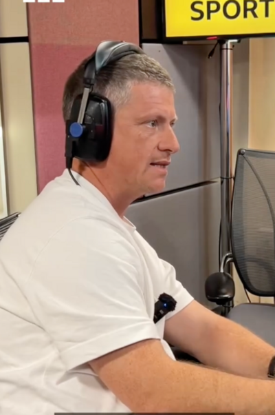 Justin Rees, wearing a white T-shirt and wearing black headphones, is in a radio studio. He looks to the right, to someone off camera.