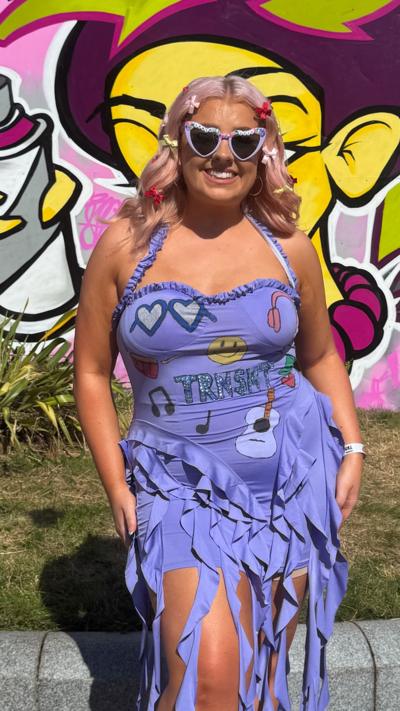 Woman wears a purple dress, sunglasses and white trainers smiles at the camera