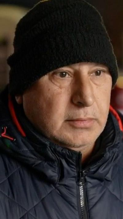 Man wearing a coat and hat stands in an empty street while being interviewed.