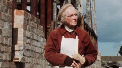 May Savidge stood in front of her partially built home, wearing work clothes and an apron, and holding a stone brick.