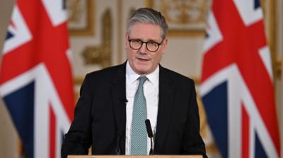 Sir Keir Starmer standing at a podium with two black microphones and two large Union Jack flags behind him