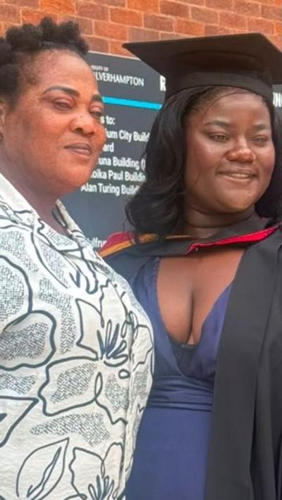 A mother and daughter at a student graduation ceremony. The mother is wearing a white patterned top. Her daughter has a blue dress, black gown and black mortar board