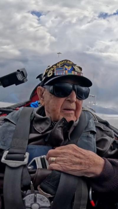 Casey Bukowski is wearing a leather jacket strapped into a plane seat, with clouds behind him and some fields visible below.