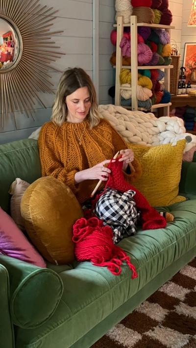 A woman sat on a green sofa knitting. She has long hair and is wearing a gold knitted top. She is using red wool