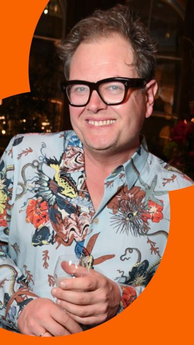 Alan Carr smiles at the camera. He is wearing a pale blue shirt with an elaborate floral pattern on it.