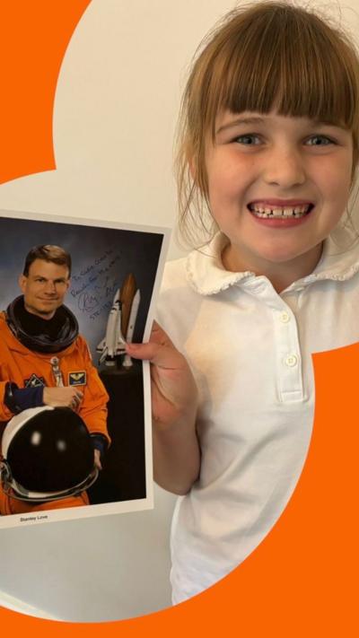 A young girl smiling, holding a picture of an astronaut in one hand and a letter in the other. She wears a white T-shirt