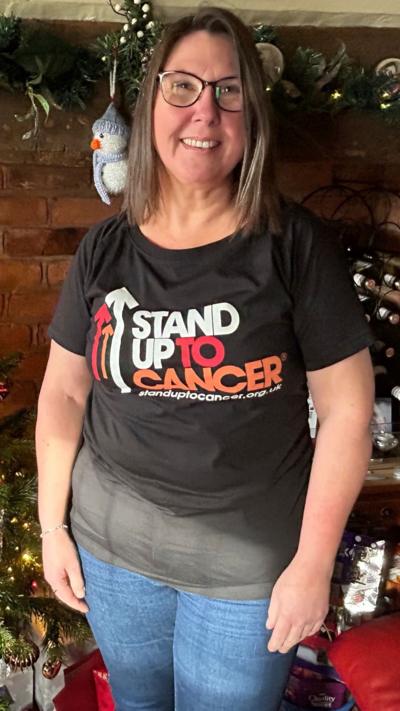 A woman with brown hair and glasses stood in front of a brick fireplace containing a Christmas tree. She is wearing blue jeans and a black t-shirt with stand up to cancer on it