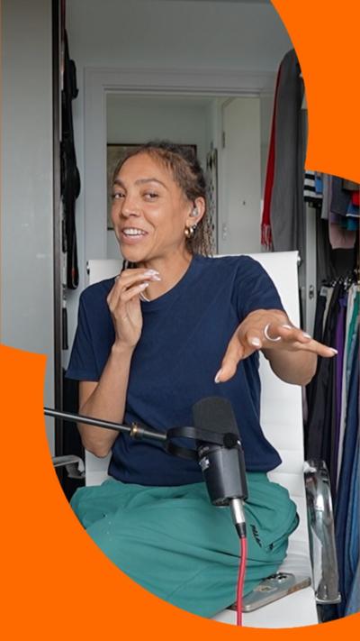 Miquita Oliver is wearing a blue top and green trousers, sat on a white chair.