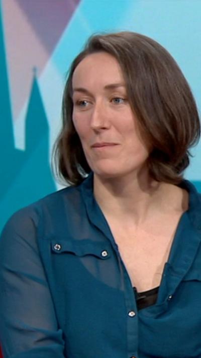 A woman is sitting in a studio wearing a blue shirt. She has brown hair.