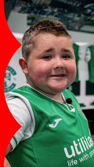 Sam Millar, eight, stands in the dressing room of Hibernian Football Club along with Dwight Gayle, one of the players. Sam is wearing a green football top and Dwight is wearing a black gilet jacket.